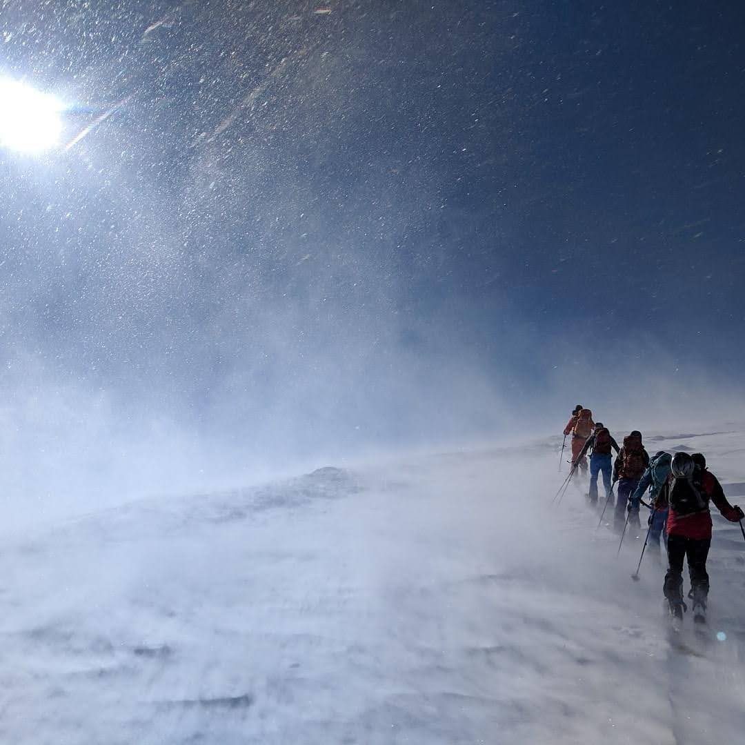 A line of backcountry skiers ascending a wind-blown ridge in bright sun and blowing snow
